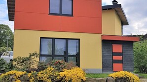 Een modern huis met twee verdiepingen, een plat dak en een schoorsteen. Op de voorgevel zijn twee ramen zichtbaar en het huis ligt aan een kinderaardig oprit.