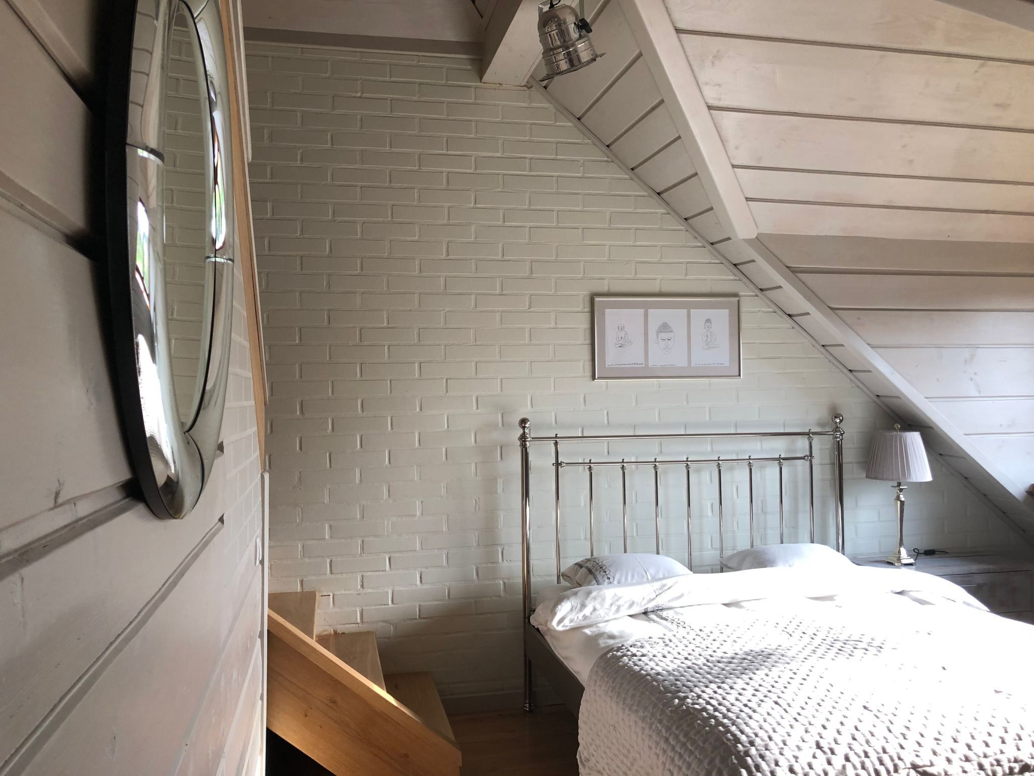 Une chambre avec un plafond en pente, un cadre de lit en métal, un mur en briques blanches et un tableau encadré au-dessus du lit.