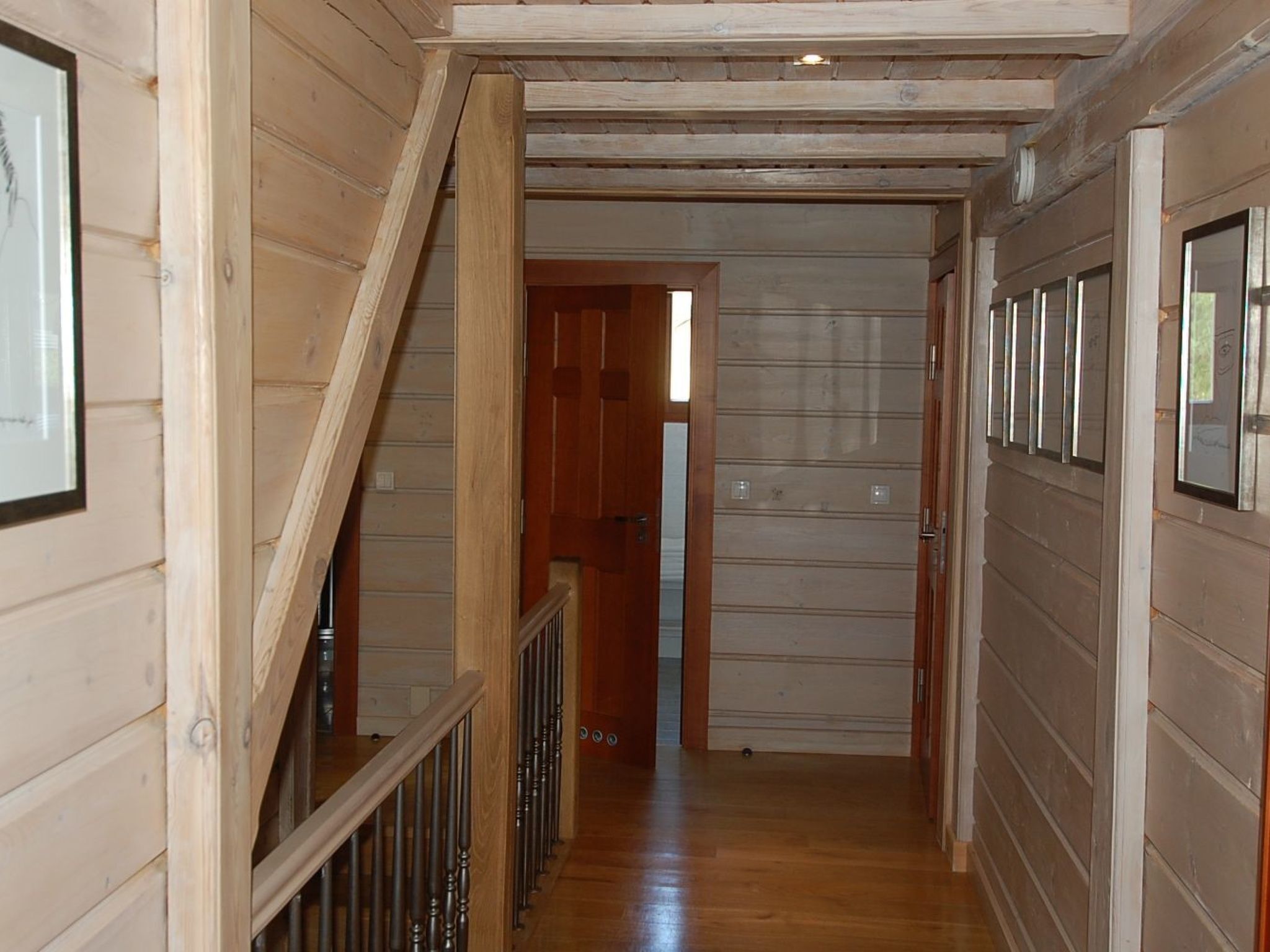 Um corredor de madeira com uma escada à esquerda, uma porta à direita e vista para outro quarto.