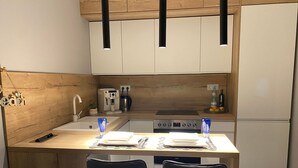 Una cocina compacta con armarios de madera, un fregadero, una cafetera y un refrigerador. Hay dos sillas en una pequeña mesa de comedor con un mantel individual y un vaso.