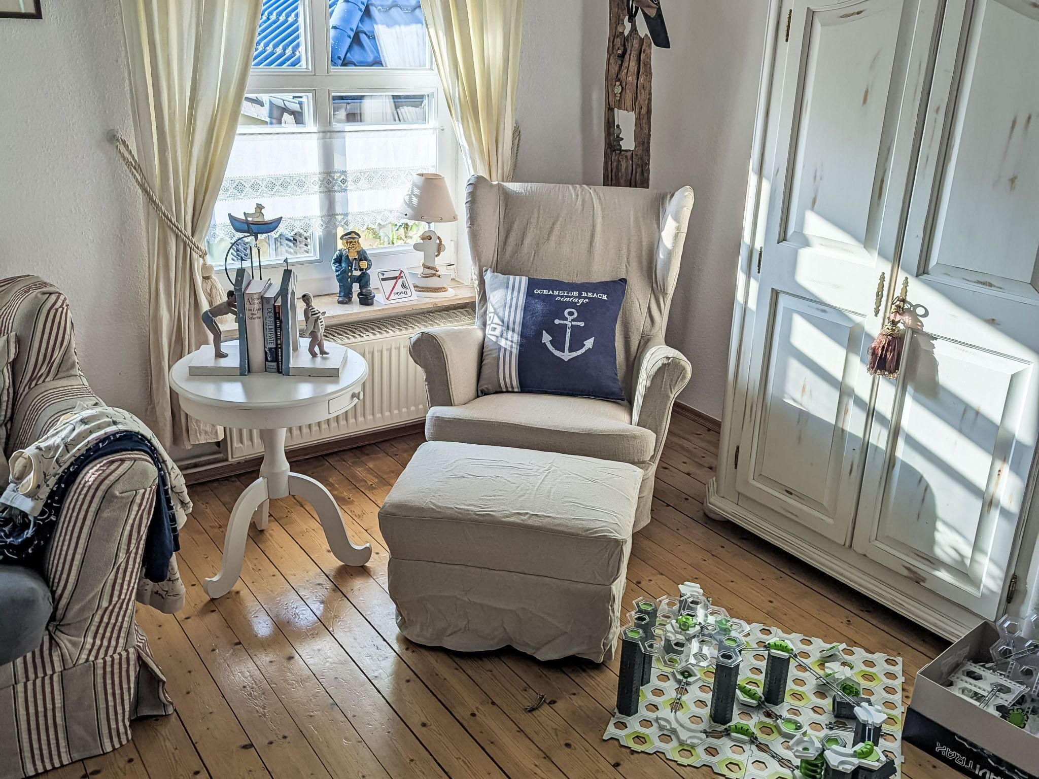 A cozy room with a striped armchair, a cushioned bench, a small table with decorative items, and a window with curtains.