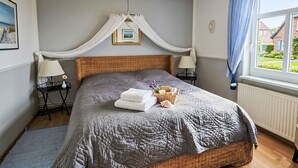 A bedroom with a large bed, a canopy, bedside tables with lamps, a wicker basket, and a view of a house through a window. - Apartment 55qm by Interhome (Ahrenshoop)