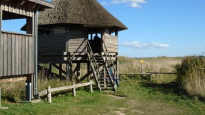 Een houten constructie met rieten dak, een trap die naar een hoger gelegen niveau leidt, en uitzicht op het omliggende landschap.