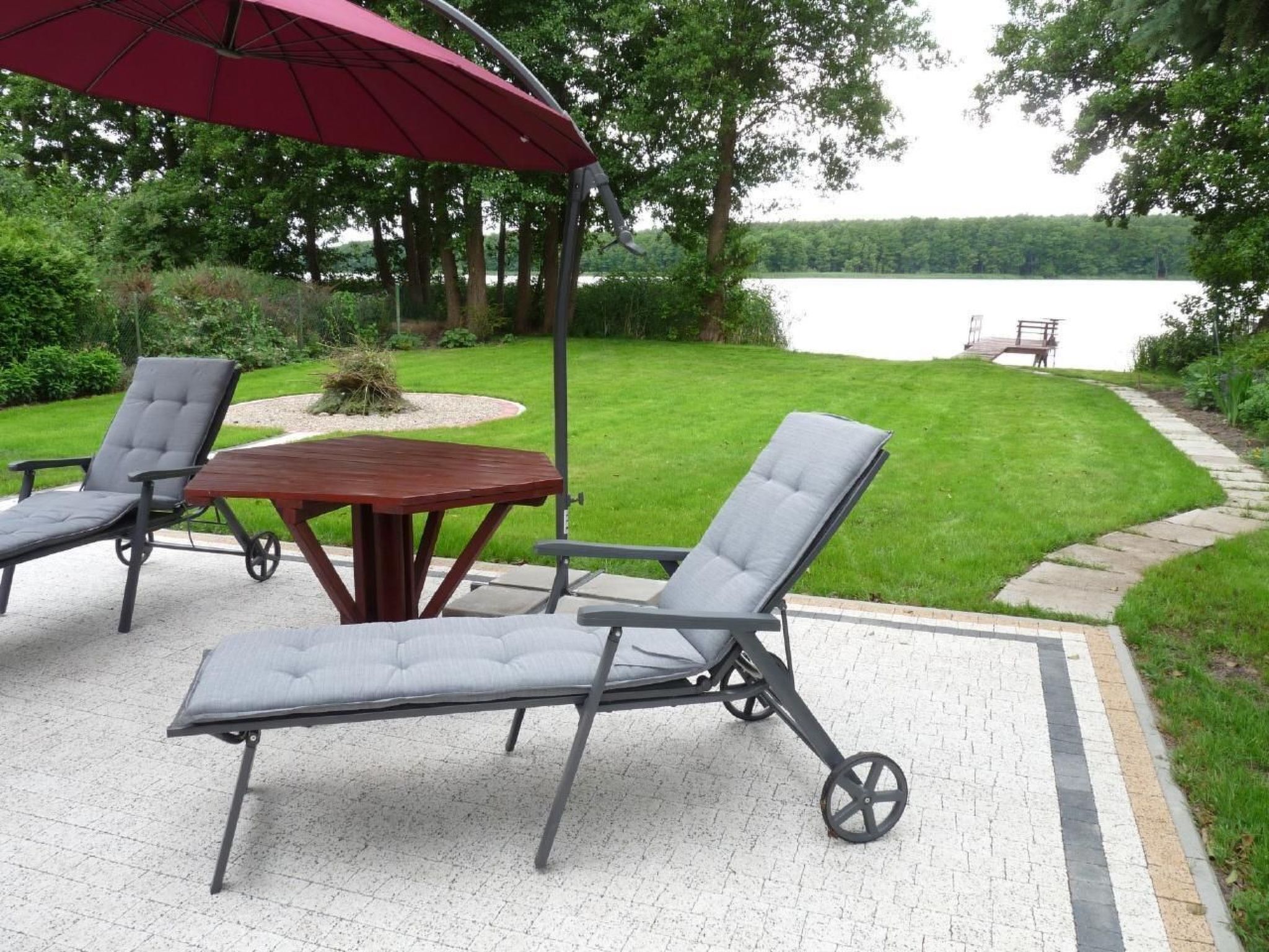 Eine Terrasse im Freien mit Tisch und zwei Lounge-Stühlen, einem roten Sonnenschirm und Blick auf einen See.