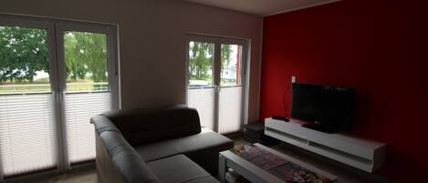 A living room with a sofa, a coffee table, a TV on a white stand, and large windows with blinds.