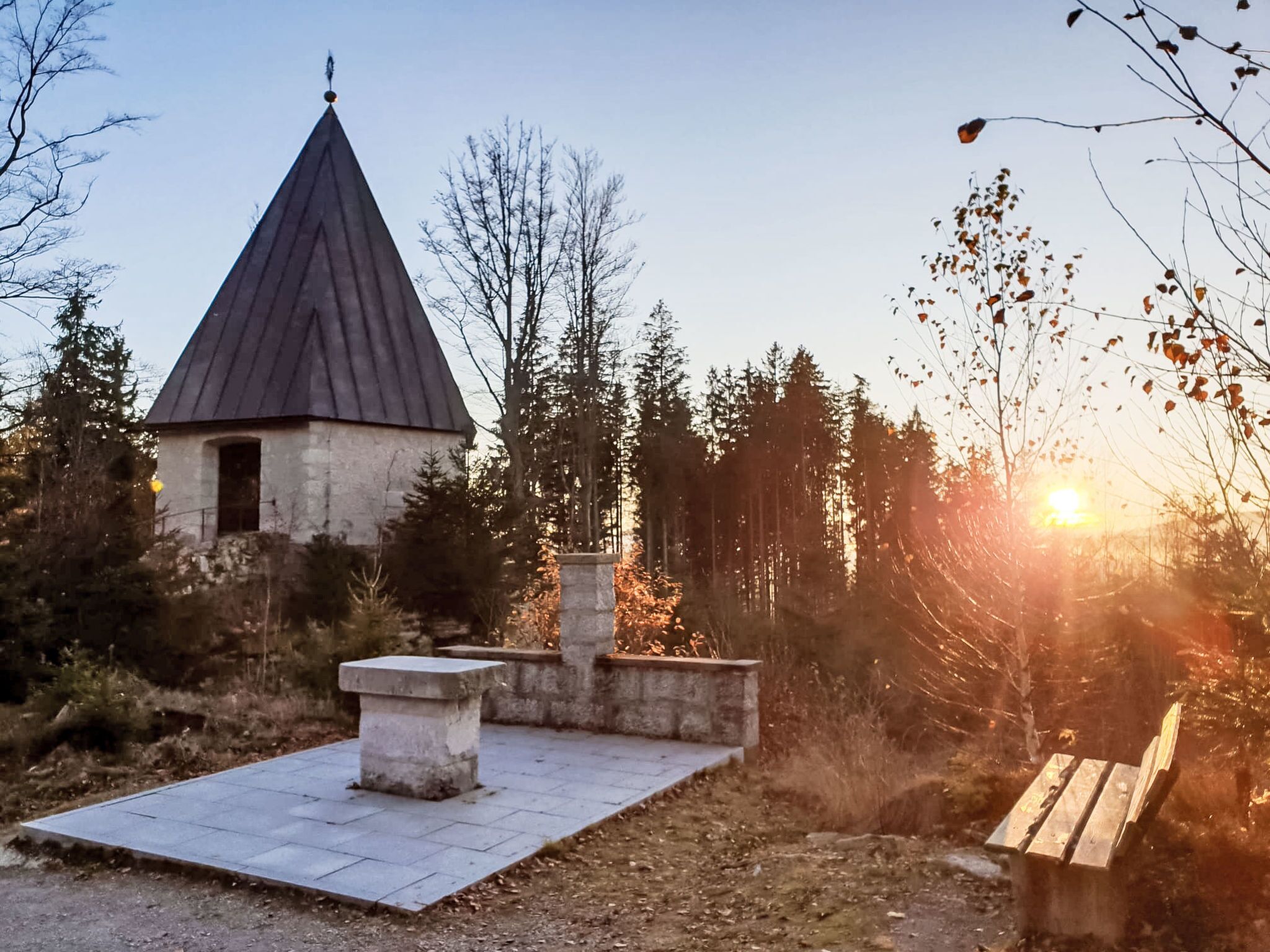 A small stone structure with a pointed roof, surrounded by trees and a bench.