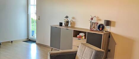 Una sala de estar con un sillón gris, un aparador de madera con objetos decorativos y un reloj en la pared.
