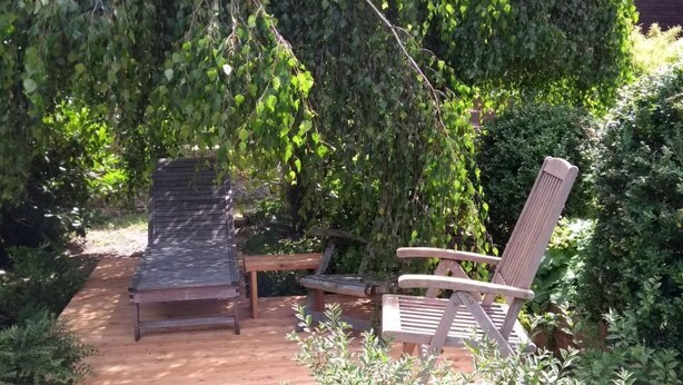Eine Holzterrasse mit zwei Holzstühlen und einem Tisch, umgeben von dichtem Grün und Bäumen.