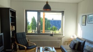 Ein Wohnzimmer mit einer Couch, einem Sessel, einem Couchtisch, einem Bücherregal und einem großen Fenster mit Blick auf Bäume und Solaranlagen.