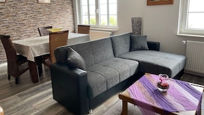 Una sala de estar con un sofá gris, una mesa de comedor con sillas de mimbre, una pared de piedra y una ventana con vistas.