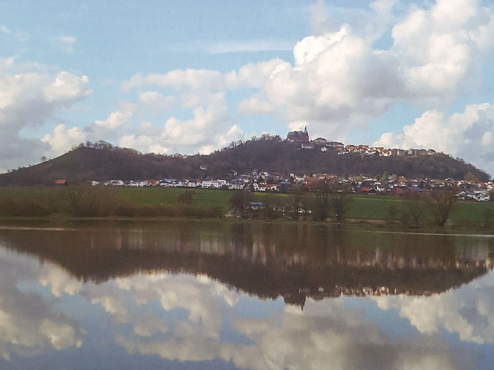 Un pueblo en la cima de una colina con una estructura similar a un castillo, rodeado de casas y una masa de agua que refleja el cielo y las nubes.