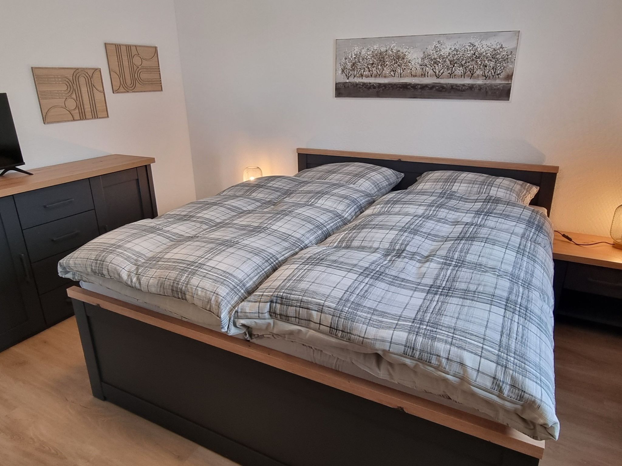 Ein Bett mit kariertem Überwurf, ein Nachttisch mit Lampe und zwei Wandbilder darüber.