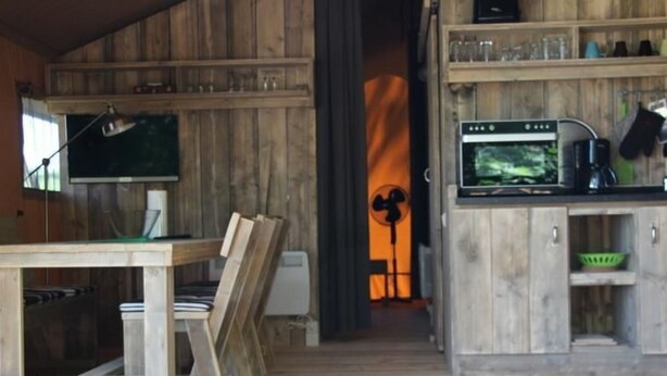 A rustic wooden interior with a dining area, a kitchen with a stove and sink, and a fan in the background. - Safarizelt in Hohenlychen mit Garten und Grill by Interhome (Lychen)