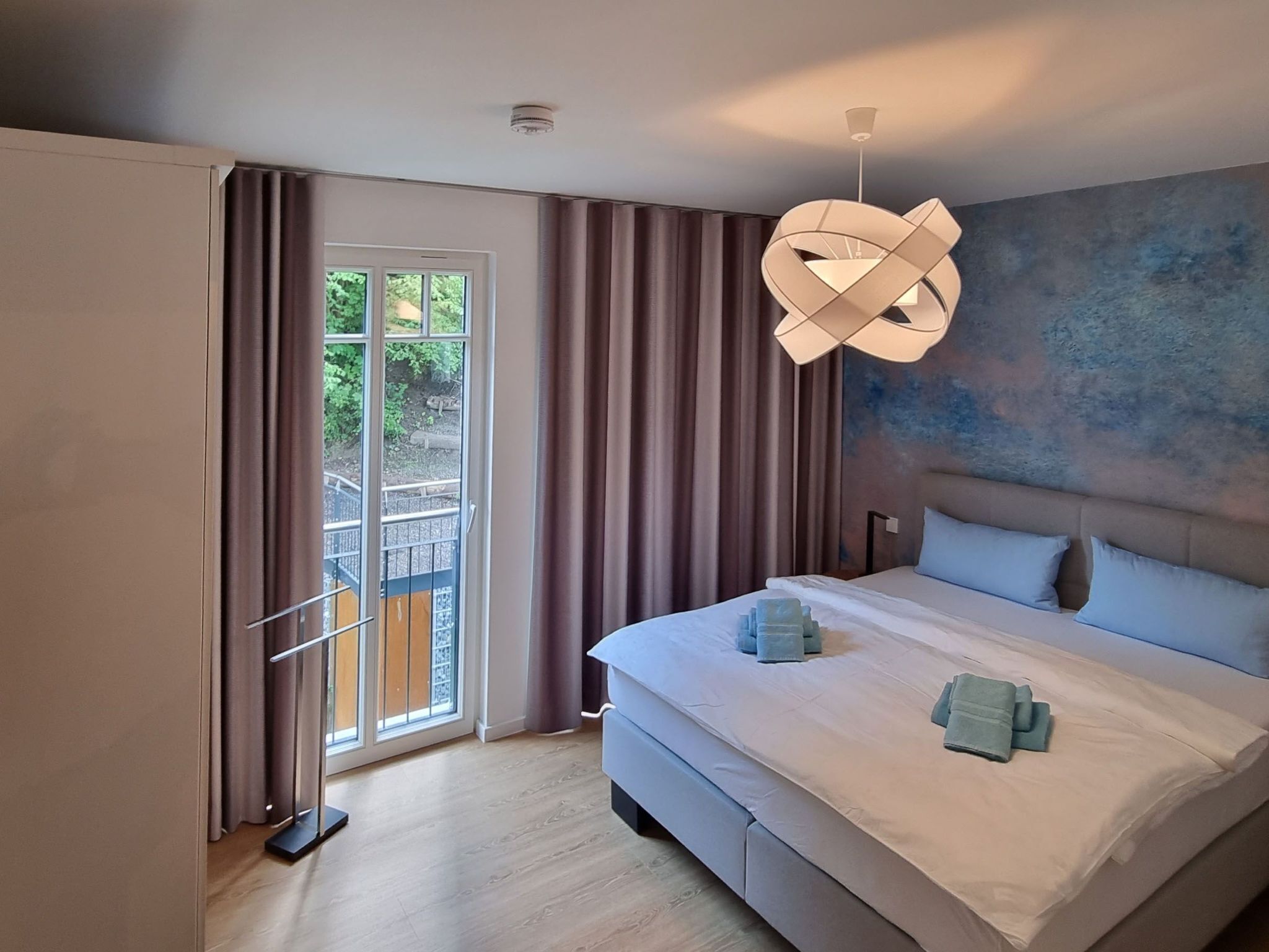 A modern hotel room with a large bed, a view through a sliding door to an outdoor area, and a unique ceiling light fixture.