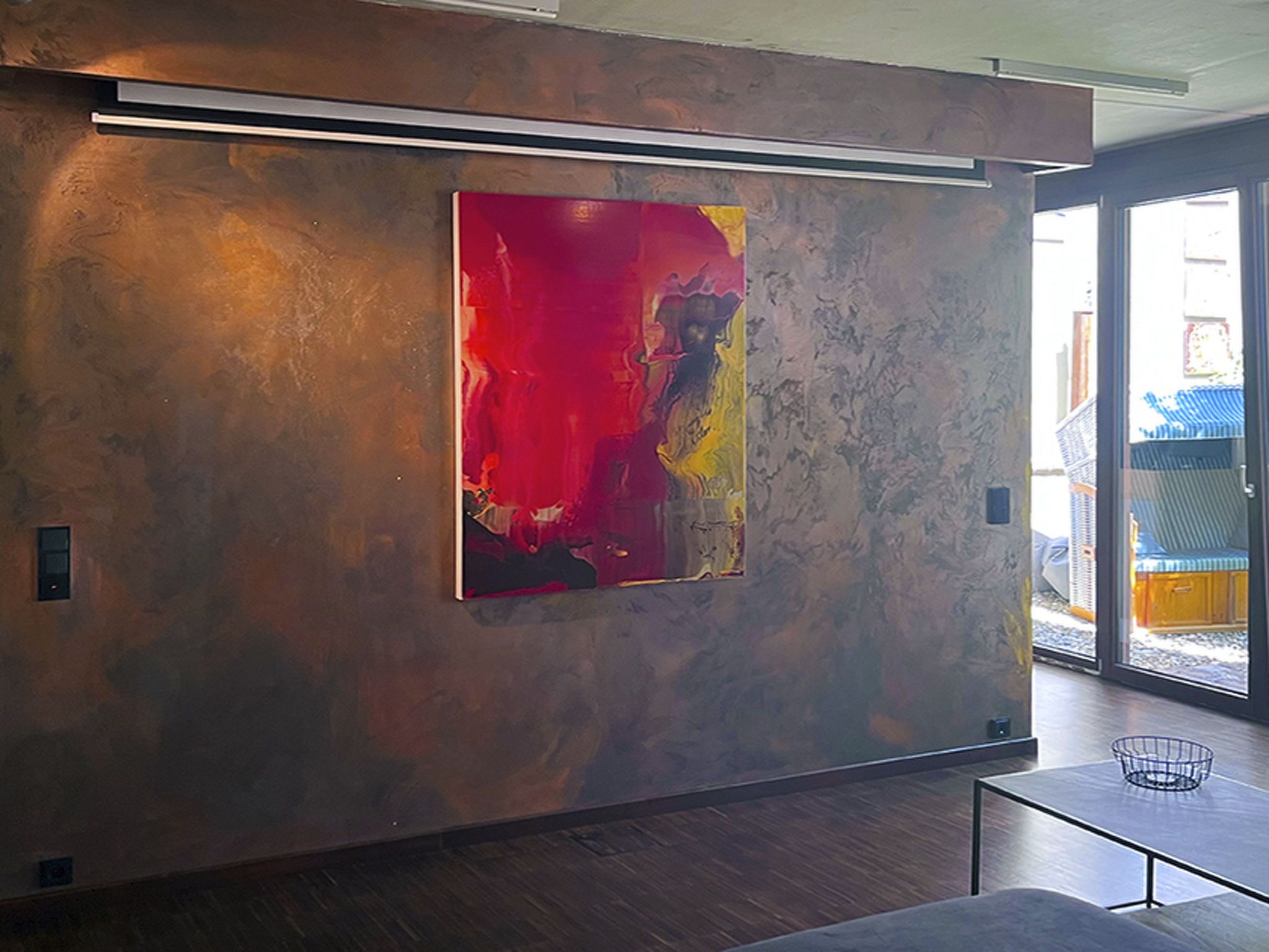 Un intérieur moderne avec un grand tableau abstrait sur un mur texturé, une porte en verre donnant sur l’extérieur et une simple table en métal.