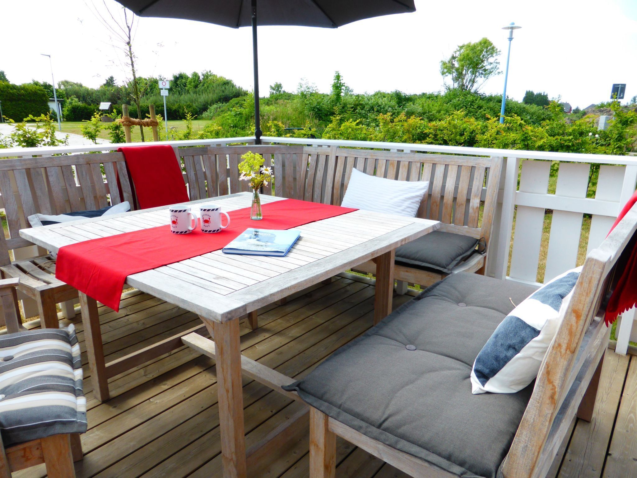 Een houten terras met een tafel en stoelen, een rode tafelkleed en een vaas met bloemen.