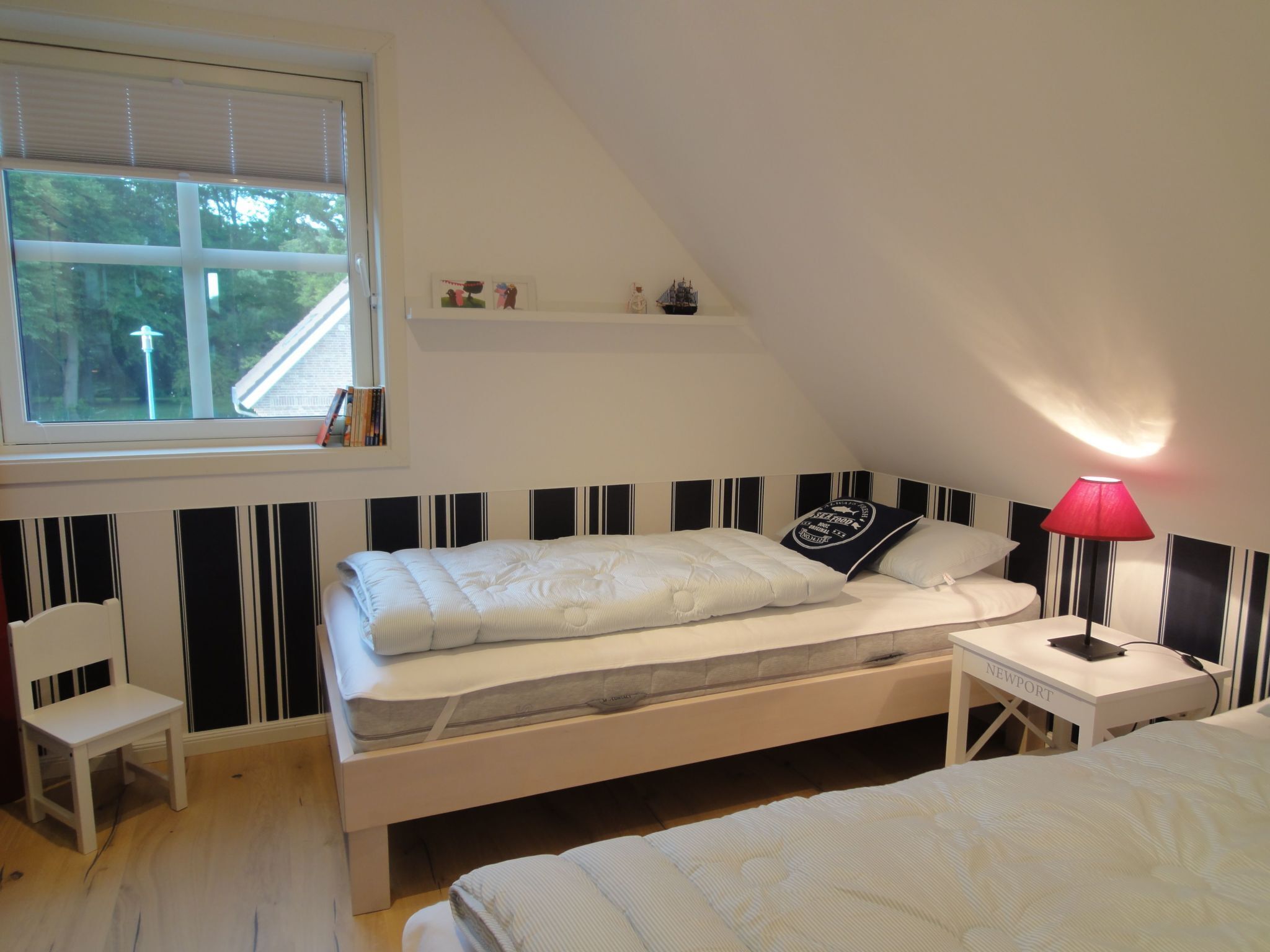 Een slaapkamer met een bed, nachtkastje, stoel, raam en een plank met kleine spullen.