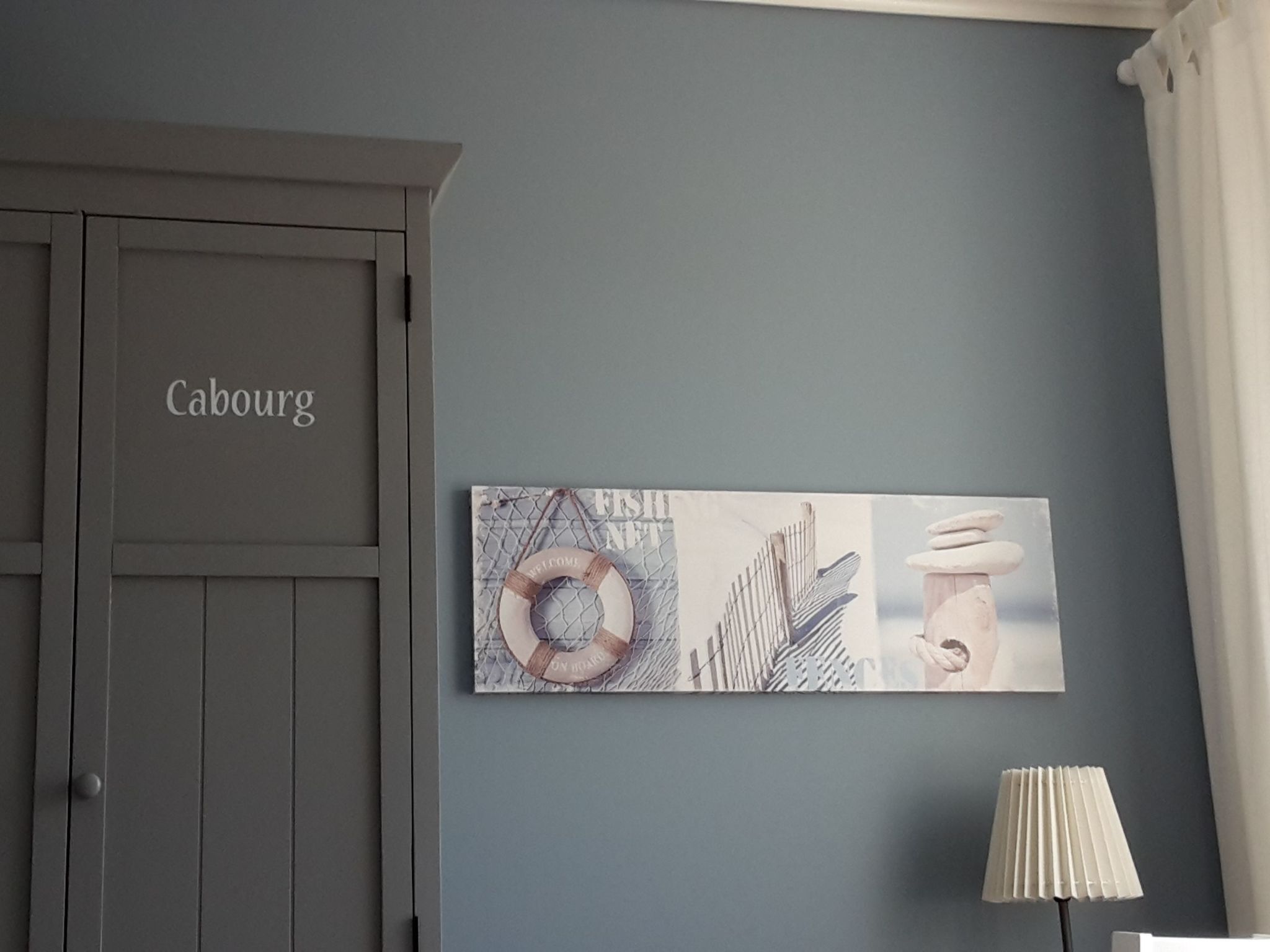 Ein Zimmer mit einer grauen Tür mit der Aufschrift 'Cabourg', einem an der Wand befestigten Kunstwerk, das einen Rettungsring und eine Hand zeigt, und einer weißen Lampe.