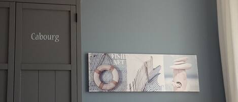 Ein Zimmer mit einer grauen Tür mit der Aufschrift 'Cabourg', einem an der Wand befestigten Kunstwerk, das einen Rettungsring und eine Hand zeigt, und einer weißen Lampe.