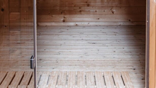 Interior de una cabaña de madera con estantes que sostienen un balde, una cuchara y una botella, todo de madera.