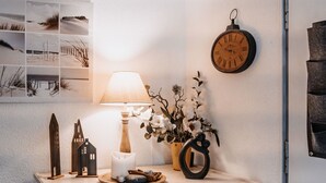 Una mesita de noche con una lámpara, un reloj, un florero con flores y una maqueta decorativa de una casa.