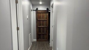 A narrow hallway with grey walls and wooden flooring, leading to a room with a large wooden door.