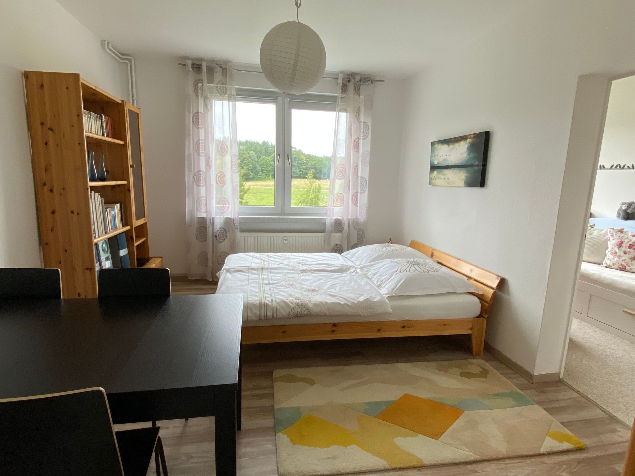 Un dormitorio con cama de madera, escritorio, estantería y vista a zonas verdes.