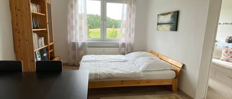 Un dormitorio con cama de madera, escritorio, estantería y vista a zonas verdes.