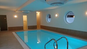 Une piscine à l’eau d’un bleu limpide, entourée d’un sol et de murs recouverts de carreaux. Des fenêtres rondes sont incrustées dans les murs, et une porte donne accès à une autre pièce.