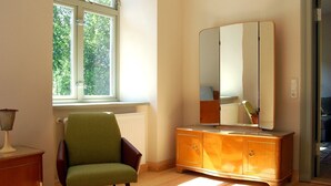 Ein Zimmer mit Holzboden, einem grünen Sessel, einem Holzschrank mit Spiegel und einem Fenster mit Blick auf Bäume.