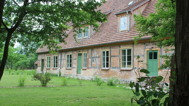 Ein traditionelles Steinhaus mit zahlreichen Fenstern und einer grünen Tür, umgeben von einer grünen Rasenfläche und Bäumen.