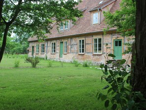 Un bâtiment traditionnel en pierre, doté de nombreuses fenêtres et d’une porte verte, entouré d’une pelouse verdoyante et d’arbres.