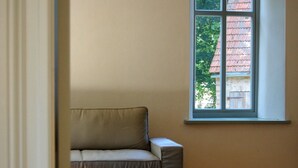 Ein Zimmer mit einem beigen Sofa, Holzboden und einem Fenster mit Blick auf ein Ziegelgebäude und Bäume.