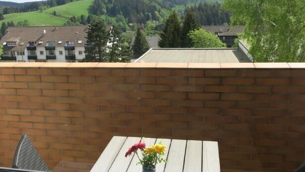 Un balcón con un muro de ladrillo, una mesa de madera con un florero y vistas a casas y colinas al fondo.