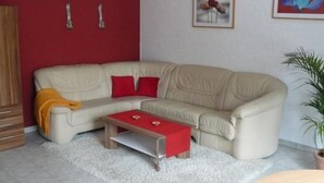 Een woonkamer met een beige bank, rode kussens, een klein houten bijzettafeltje en een crèmekleurig shagtapijt.