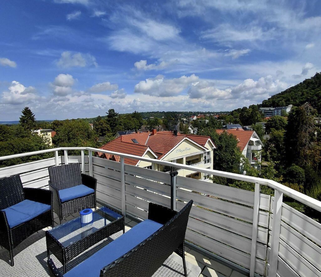 Un balcon doté de meubles en osier et d’une table, offrant une vue sur un quartier résidentiel avec ses maisons et sa verdure.