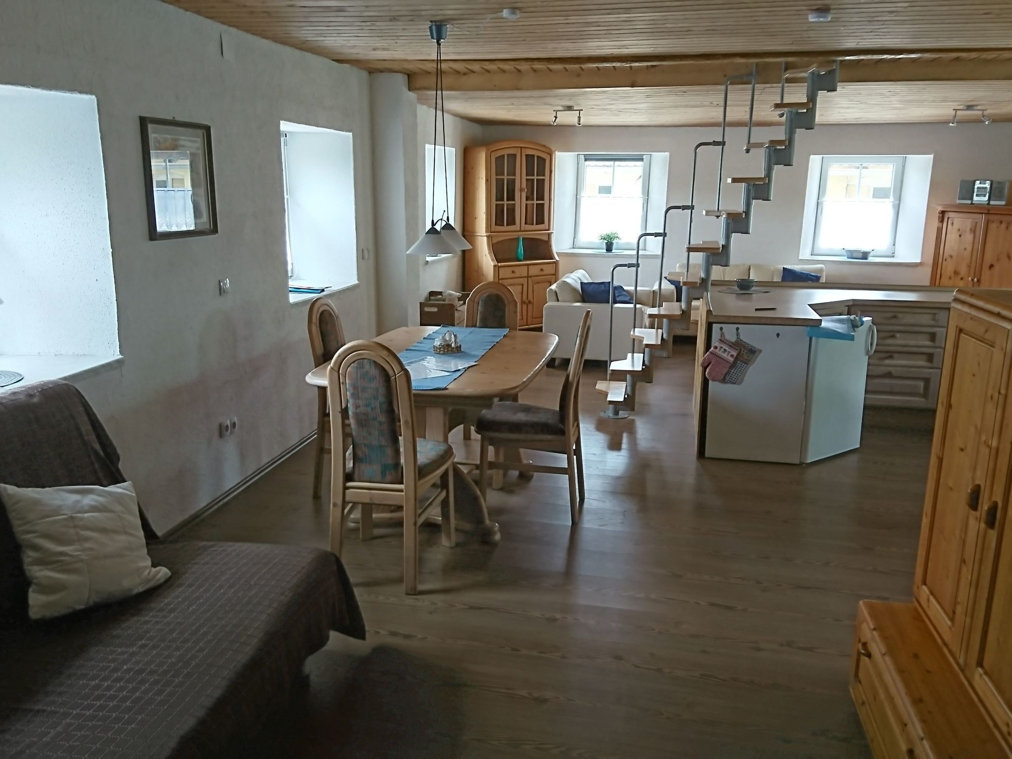 A spacious living area with a dining table, chairs, a sofa, a kitchen area with a refrigerator, and a wooden staircase.