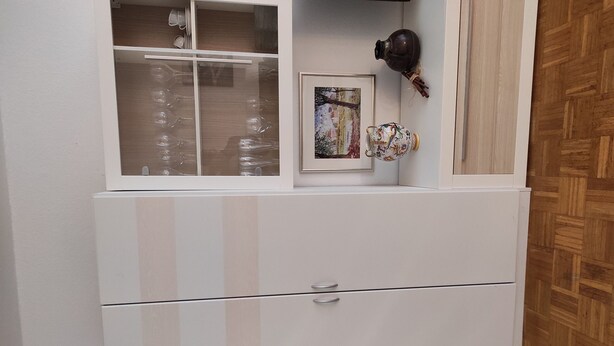 Un meuble blanc, avec une partie supérieure dont la façade est en verre, contenant de la verrerie et un tableau encadré.