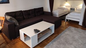 Un salon moderne avec un canapé marron foncé, une table basse blanche, un lit blanc et une petite table de chevet avec une lampe.