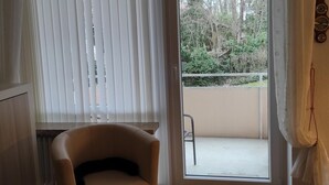 Une pièce dotée d’un parquet en bois, d’un fauteuil beige, d’une porte-fenêtre coulissante donnant sur un espace extérieur avec un fauteuil, et d’une horloge murale.