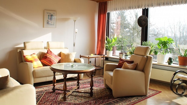 Een woonkamer met een bank, fauteuil en salontafel. Op de vensterbank staan planten en op de vloer ligt een vloerkleed.