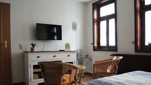 Une chambre à coucher avec un lit, deux fauteuils en osier, un téléviseur posé sur une commode blanche et une fenêtre donnant sur un paysage agréable.