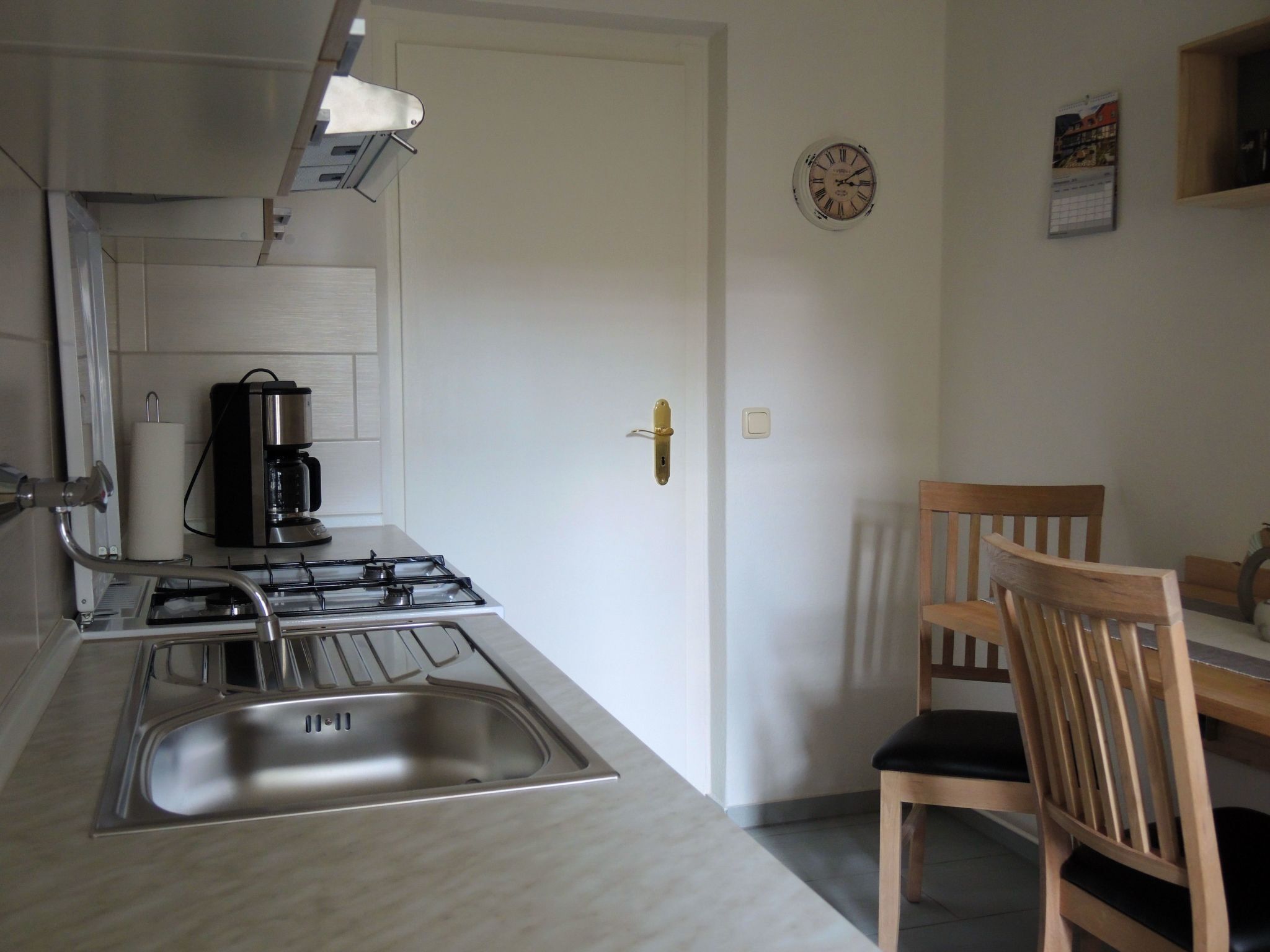 A kitchen with a gas stove, a coffee maker, and a sink. There is a wooden chair and a small table with a clock on the wall.