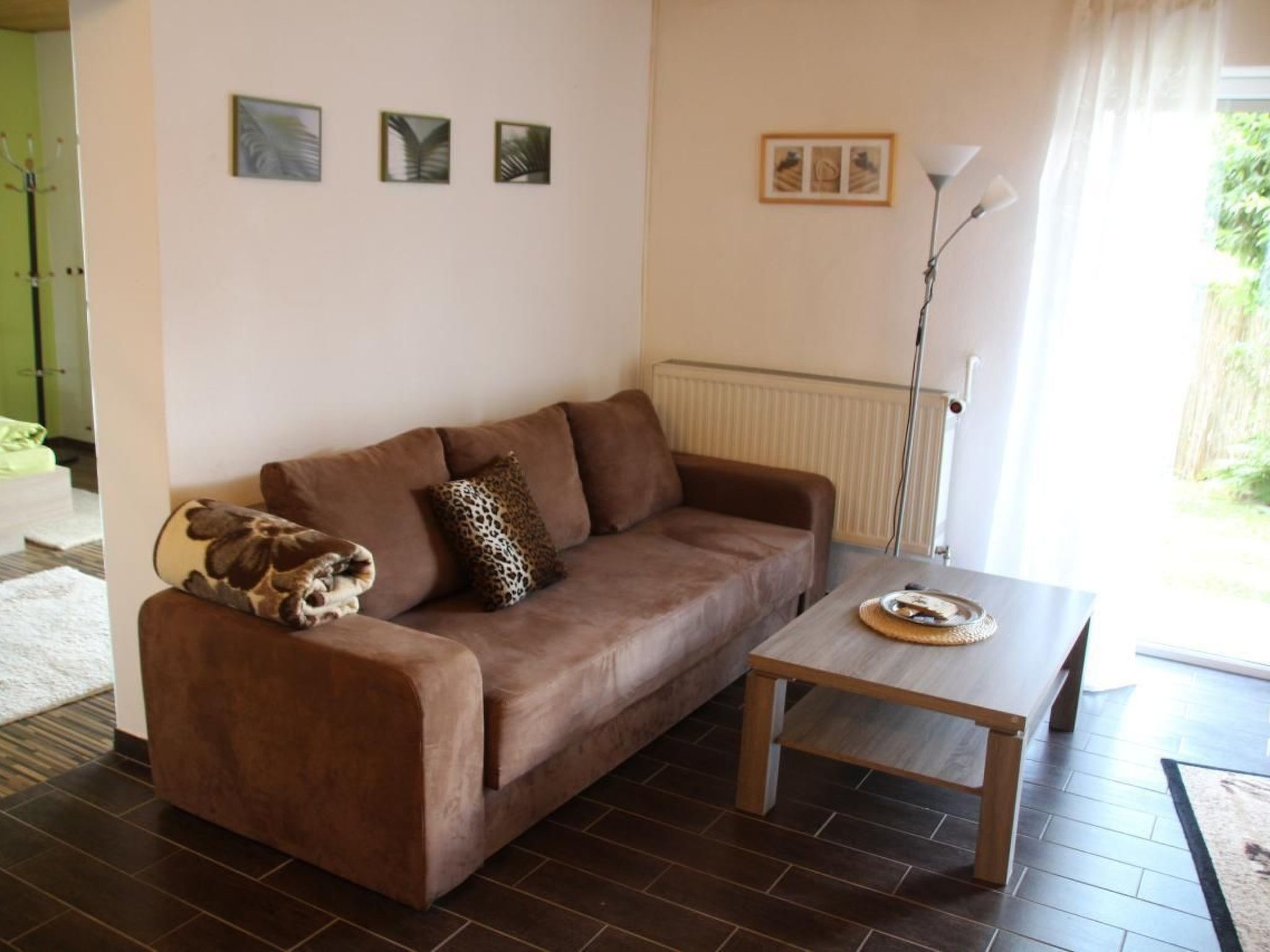 A living room with a brown sofa, a wooden coffee table, and a leopard print pillow. There are three framed pictures on the wall and a floor lamp.