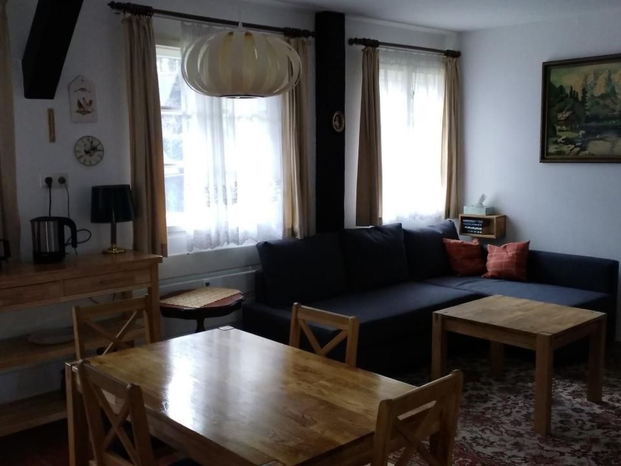 Ein gemütliches Wohnzimmer mit einem Holzesstisch, einem dunklen Sofa und einem kleinen Beistelltisch. Ein Fenster mit Vorhängen, eine Lampe und ein Bild an der Wand sind zu sehen.