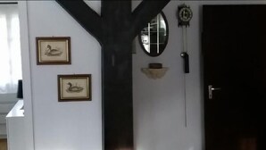 Une pièce avec un plancher en bois foncé, un mur blanc orné de deux tableaux encadrés, un miroir rond, une horloge murale et une porte.