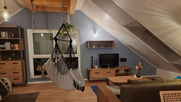 Un ambient de saló estufa, d'estil loft, amb una cadira enfilada, mobles de fusta, un televisor i una biblioteca.