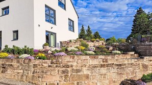 Edifici blanc amb nombroses finestres, mur de pedra amb parterres de flors i un cel serè.