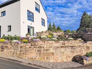 A white building with multiple windows, a stone retaining wall with flower beds, and a clear sky. - Panoramadomizil by Interhome (Goldkronach)