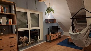 Habitació acollidora amb terra de fusta, llit amb llençol estampat, prestatgeries, televisor sobre un suport de fusta, planta penjant i cadira de relax.